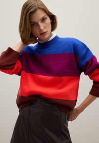Bunter Strickpullover mit horizontalen Streifen in Blau, Lila, Rot und Braun. Rippbündchen an den Ärmeln und am Kragen. Kombiniert mit einem schwarzen Rock.