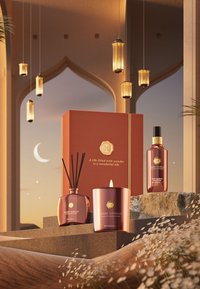 Samling av Suede Vanilla-produkter: et kobberfarget stearinlys med flamme, en diffuser og en spray, plassert mot en dekorativ boks og stein.