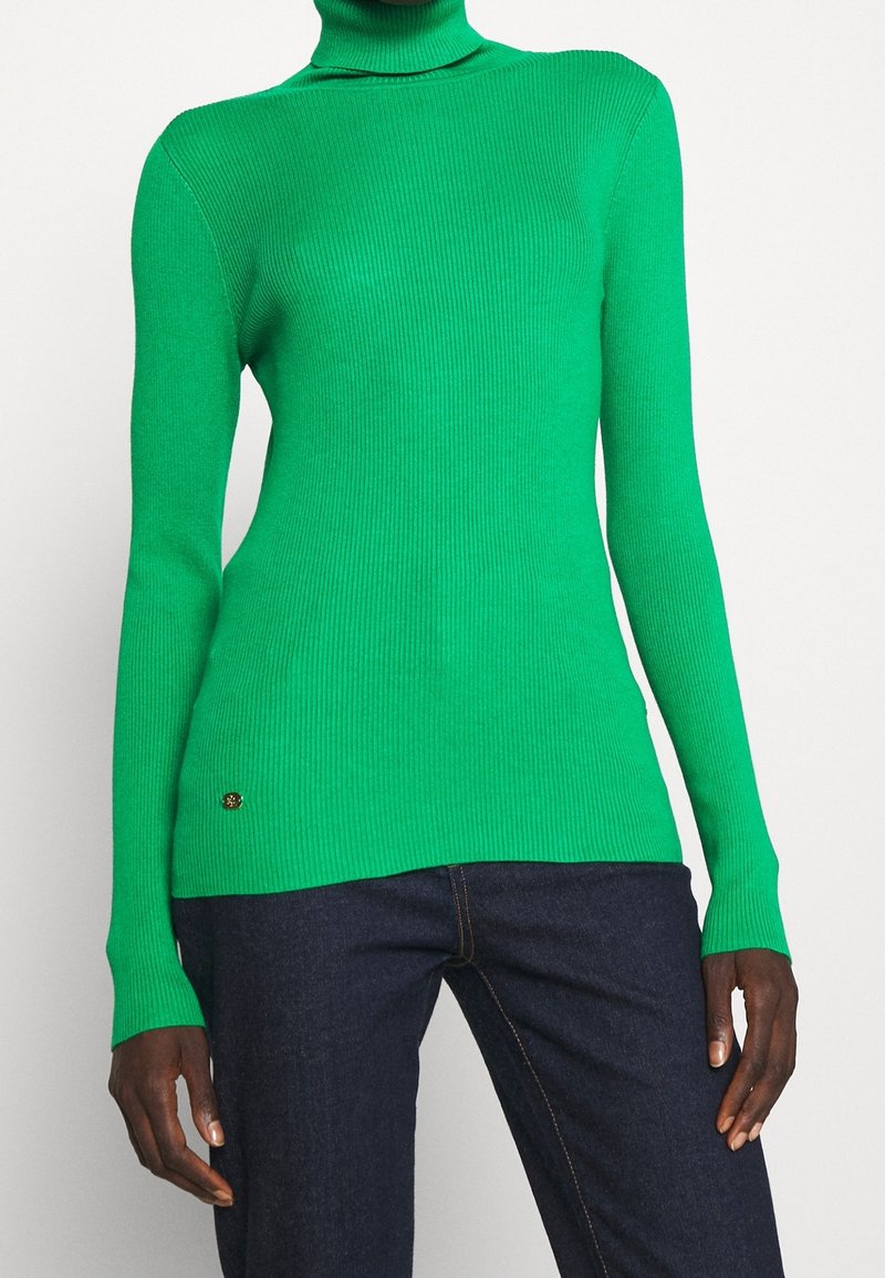 Personne portant un pull à col roulé côtelé vert vif à manches longues et un jean bleu foncé, debout devant un fond uni.