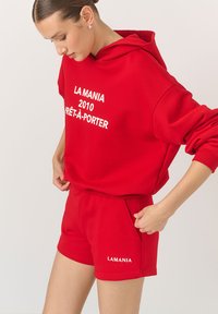 Czerwony zestaw z kapturem i szortami wykonany z bawełny, z białym nadrukowanym tekstem. Bluza z kapturem zawiera sznurek do regulacji i kieszeń typu kangur. Szorty mają kieszenie boczne.