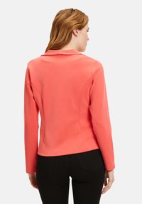 Korallfarbene taillierte Jacke mit Schalkragen, langen Ärmeln und glatter Textur. Verfügt über eine Nahtdetailierung und einen geschichteten Saum. Kombiniert mit schwarzen Hosen.