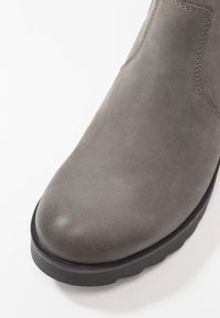 Gros plan sur une botte cheville en daim gris avec semelle en caoutchouc noir et couture apparente sur la partie supérieure.