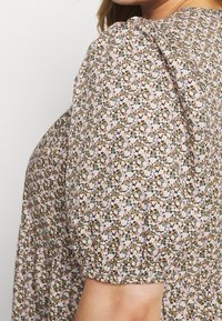 Kobieta w sukience z bufiastymi rękawami w mały kwiatowy wzór w kolorach różowym, białym, zielonym i musztardowym.