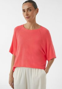 Korale dzianinowa bluzka z krótkim rękawem, okrągłym dekoltem i szczegółami z otwartego splotu na ramionach, zestawiona z jasnymi, zwiewnymi spodniami.