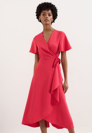 Mujer con un vestido cruzado rojo con mangas vuelo y detalle de cintura anudada, de pie frente a un fondo claro y liso.