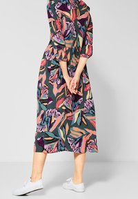 Šaty s květinovým potiskem, které mají barevné listy v zelených, růžových a fialových tónech. Šaty mají dlouhé rukávy a vrstvený design, doplněné o bílé tenisky.