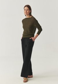 Olijfgroen gebreid sweater met lange mouwen, gecombineerd met zwarte losse broek. Lichte textuur zichtbaar, model draagt tan schuifsandalen.