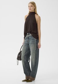 Jeune femme portant un top noir sans manches à col montant, un jean large délavé avec des revers de manches, des talons pointus olive, une ceinture chaîne argentée, portant un sac à main noir.