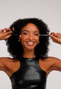 Czarna, bezrękawnikowa bluzka na uśmiechniętej osobie trzymającej dwa czarne ołówki do makijażu, z błyszczącym wykończeniem i kręconymi włosami.