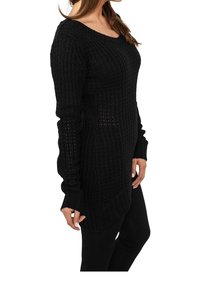 Maglione nero a maglia con un motivo testurizzato, scollo aperto e maniche lunghe, abbinato a leggings neri. Presenta una vestibilità comoda e una leggera asimmetria.
