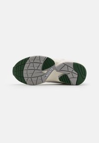 Lacoste tenisica s crnom i zelenom gumenom potplatom, teksturiranim uzorkom, i sivim međupotplatom s reljefnim logotipom.