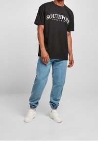 Camiseta de algodón negra con el texto blanco "SOUTHPOLE ORIGINAL", combinada con joggers de mezclilla azul claro y zapatillas blancas.