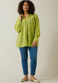 Camicia di cotone verde con motivi ad occhiello, maniche lunghe e colletto. Abbinata a jeans skinny blu e a scarpe ballerine dorate.