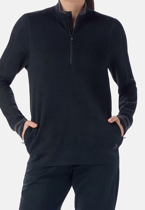 Kvinde iført en sort kvart-lynlås trøje med hænderne i forlommerne, kombineret med sorte bukser, mod en ensfarvet baggrund.