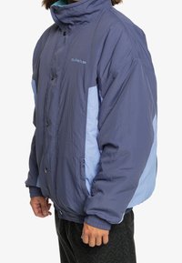 Personne portant une doudoune Quiksilver bicolore bleue avec boutons-pression, poignets élastiques et poches latérales zippées, debout de profil.