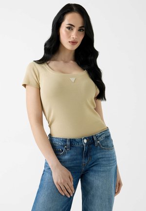 Jeune femme aux longs cheveux noirs portant un haut beige à manches courtes et un jean bleu, debout devant un fond blanc uni.