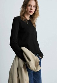 Maglietta nera a maniche lunghe in tessuto morbido, abbinata a una giacca esterna beige chiaro e jeans blu. La maglietta presenta un collo rotondo.