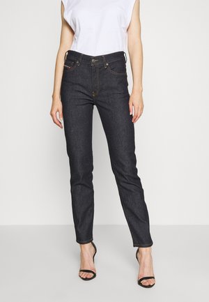 Personne portant un jean slim foncé, un haut blanc sans manches et des sandales noires à talons ouverts, debout devant un fond uni.