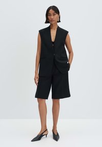 Schwarzer ärmelloser Blazer mit strukturierten Schultern, zwei Taschen, kombiniert mit maßgeschneiderten schwarzen Shorts, getragen mit schwarzen Slingback-Absätzen.