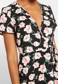 Wrap dress in a black fabric featuring pink floral prints. Short sleeves, v-neckline, adjustable tie at the waist, smooth texture.