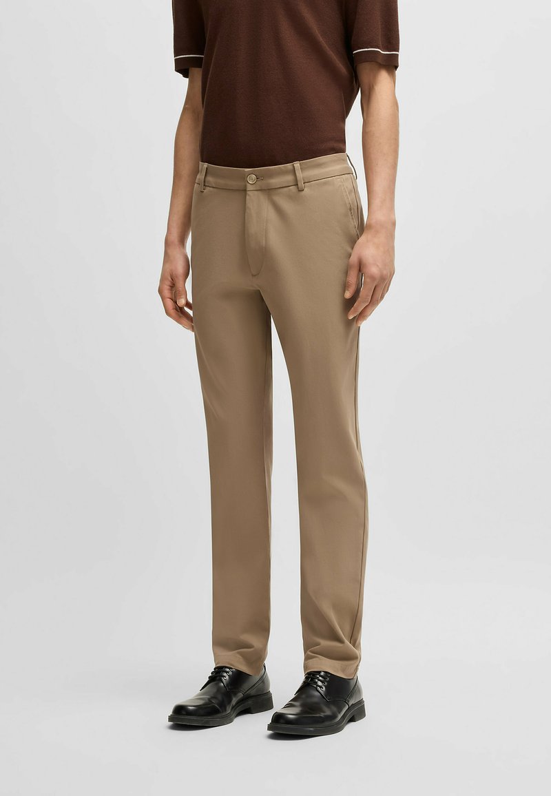 Beżowe spodnie wykonane z gładkiej tkaniny, o prostym fasonie nogawki, z zasuwanymi z przodu guzikami i bocznymi kieszeniami, w zestawie z czarnymi butami.