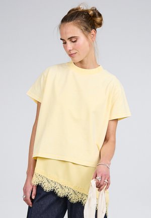 Joven con peinado de doble moño lleva blusas amarillas superpuestas con borde de encaje y vaqueros oscuros, sosteniendo una bufanda de color claro en la mano derecha.