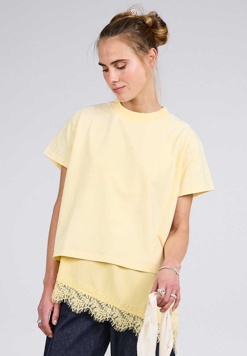 Mladá žena s dvojitým účesem nosí vrstvené žluté topy s krajkovým lemem a tmavé džíny, v pravé ruce drží světle zbarvený šátek.