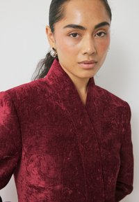 Karmino aksominė suknelė su aukšta apykakle ir tekstūruotu paviršiumi, turinti lygų apdailą ir struktūruotus pečius. Sidabro auskarai suteikia detalizacijos.