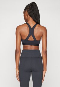 Γυναίκα με πλεξούδες, φορώντας μαύρο sports bra με racerback και ψηλόμεσα σκούρα γκρίζα κολάν, στέκεται με την πλάτη στραμμένη.
