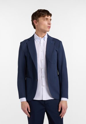 Jonge man die een marineblauw colbert en wit overhemd draagt, kijkt opzij tegen een effen witte achtergrond.