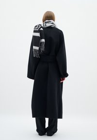 Lungo cappotto nero con vita cinturata, abbinato a una sciarpa a righe bianche e nere. Tessuto morbido, design dritto e lunghezza alla caviglia.