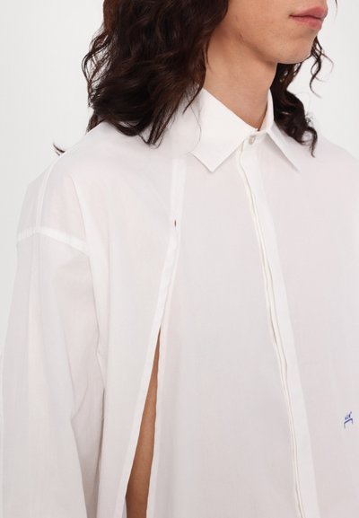 Chemise blanche à boutons avec un design partiel, fabriquée en tissu léger. Dotée d'un col classique et d'un détail brodé bleu sur la poitrine.
