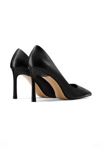 Un paio di scarpe nere in pelle con punta affusolata e tacchi a stiletto, viste da un angolo laterale-posteriore su uno sfondo bianco.