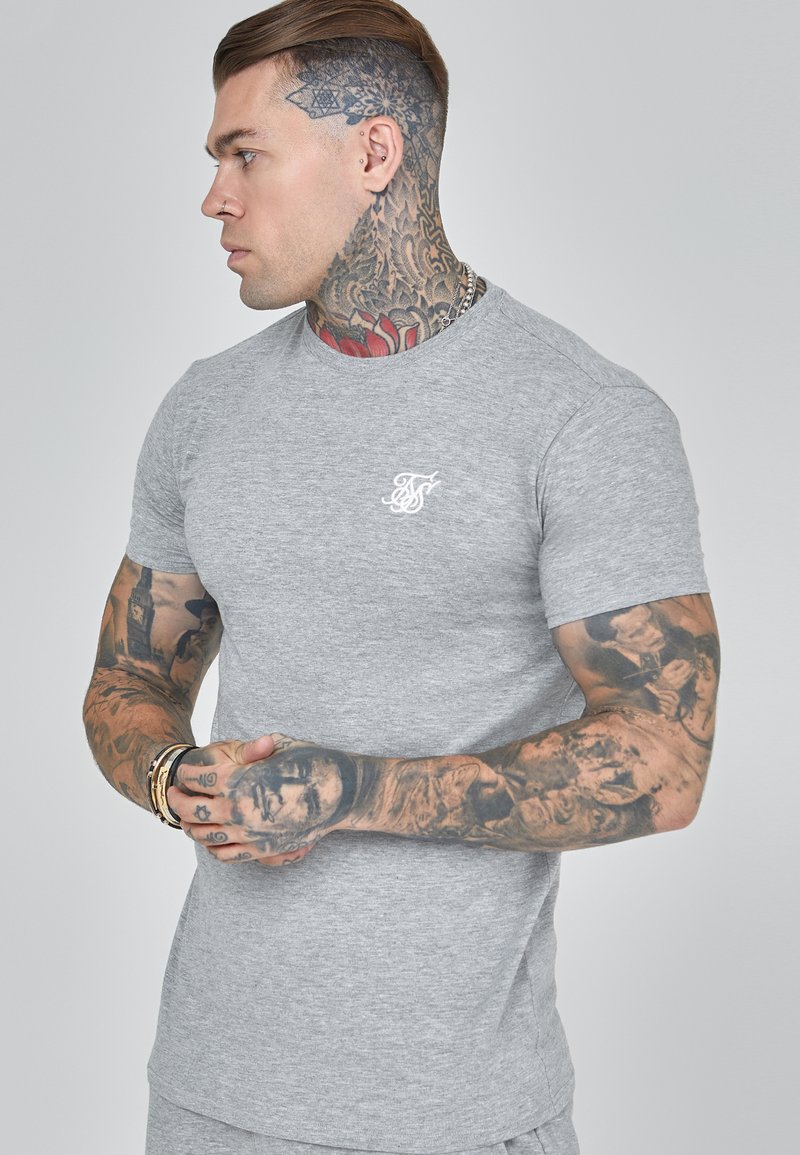 Grijze katoenen T-shirt met een ronde hals, korte mouwen en een wit geborduurd logo op de borst. Bevat intricates tattoo-details op de armen.