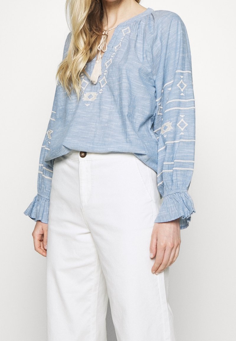 Femme portant un chemisier bleu clair brodé à manches longues avec un lien à pompons et un pantalon blanc taille haute à jambes larges.