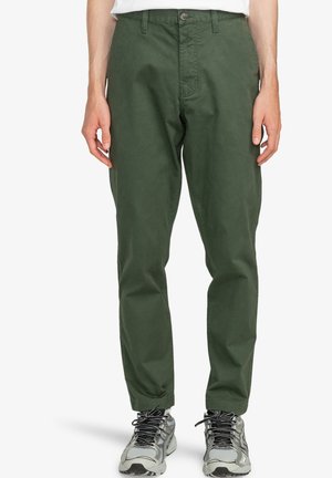Pantalones de algodón de color verde oscuro con un diseño de pierna recta, cierre frontal de botón y bolsillos laterales, combinados con zapatillas deportivas grises.