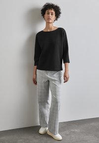 Zwarte gebreide trui met een brede halslijn, gecombineerd met lichtgrijze geruite wijde broekspijpen. Het model draagt witte sneakers. Eenvoudige achtergrond.