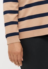 Left hand of person wearing beige sweater with navy stripes and black pants against a plain light background.