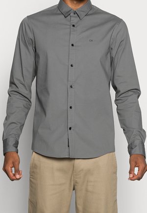Homme portant une chemise grise ajustée à manches longues avec des boutons noirs et un pantalon beige, debout les bras légèrement pliés.