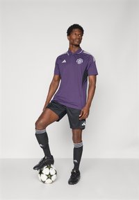 Lilla polo skjorte med sorte accenter, tre striber på skuldrene, Manchester United logo, parret med sorte shorts og fodboldstrømper, stående på en bold.