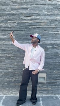 Egy világos rózsaszín gombos ingben, fekete nadrágban és farmer sapkában álló személy, aki telefont tart szürke kőfal mellett.