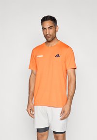 Πορτοκαλί αθλητική μπλούζα με στρογγυλή λαιμόκοψη, με λογότυπο "TENNIS" και μπλε-σκούρες ρίγες Adidas. Φορεμένη με λευκά σορτς και γκρι εσώρουχα.