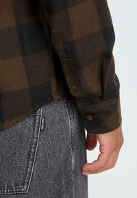 Bruin en zwart geruite flanellenshirt met knoopmanchetten, gedragen over grijze jeans met een zichtbaar label, hand rustend aan de zijkant.
