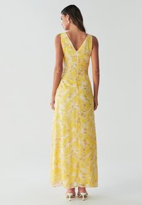 Maxi šaty zo žltého šifónu s kvetinovým vzorom. Majú výstrih do V na chrbte, bez rukávov a jemný lesk. Na zadnej strane zapínanie na zips.