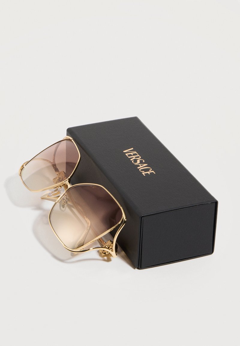Lunettes de soleil teintées à monture dorée avec des verres dégradés posées à côté d'une boîte noire Versace sur une surface blanche.