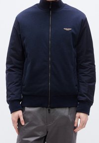 Homme portant une veste zippée bleu marine foncé avec des poignets côtelés et un petit emblème ailé sur la poitrine, associée à un pantalon gris.