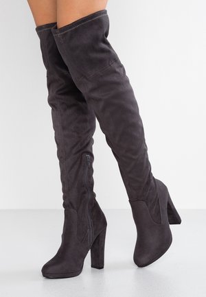 Schwarze Overknee-Stiefel aus Wildleder mit Blockabsätzen und seitlichen Reißverschlüssen, getragen an überkreuzten Beinen vor einfachem Hintergrund.