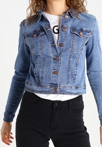 Chaqueta de mezclilla azul claro con un diseño recortado, dos bolsillos en el pecho, botones de bronce y una textura ligeramente deslavada. Llevada sobre una camisa blanca.