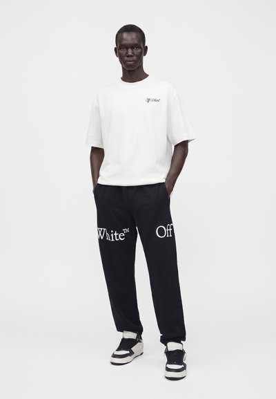 Homme portant un t-shirt blanc Off-White et un pantalon de jogging noir avec un grand texte Off-White, debout les mains dans les poches sur un fond uni.