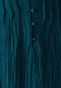 Tyrkysová texturovaná látka s vertikálními sklady; obsahuje čtyři kulaté, texturované knoflíky podél středu. Měkký, pomačkaný vzhled.